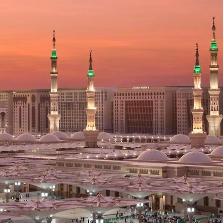 Intercontinental Dar Al Iman Madinah By Ihg Hotel Medina Exterior foto The photo depicts a scenic view of a mosque complex, likely during sunset, with several minarets illuminated and showcasing green lights. The architecture features domed roofs, and the setting includes a backdrop of buildings, all bathed in a warm, o