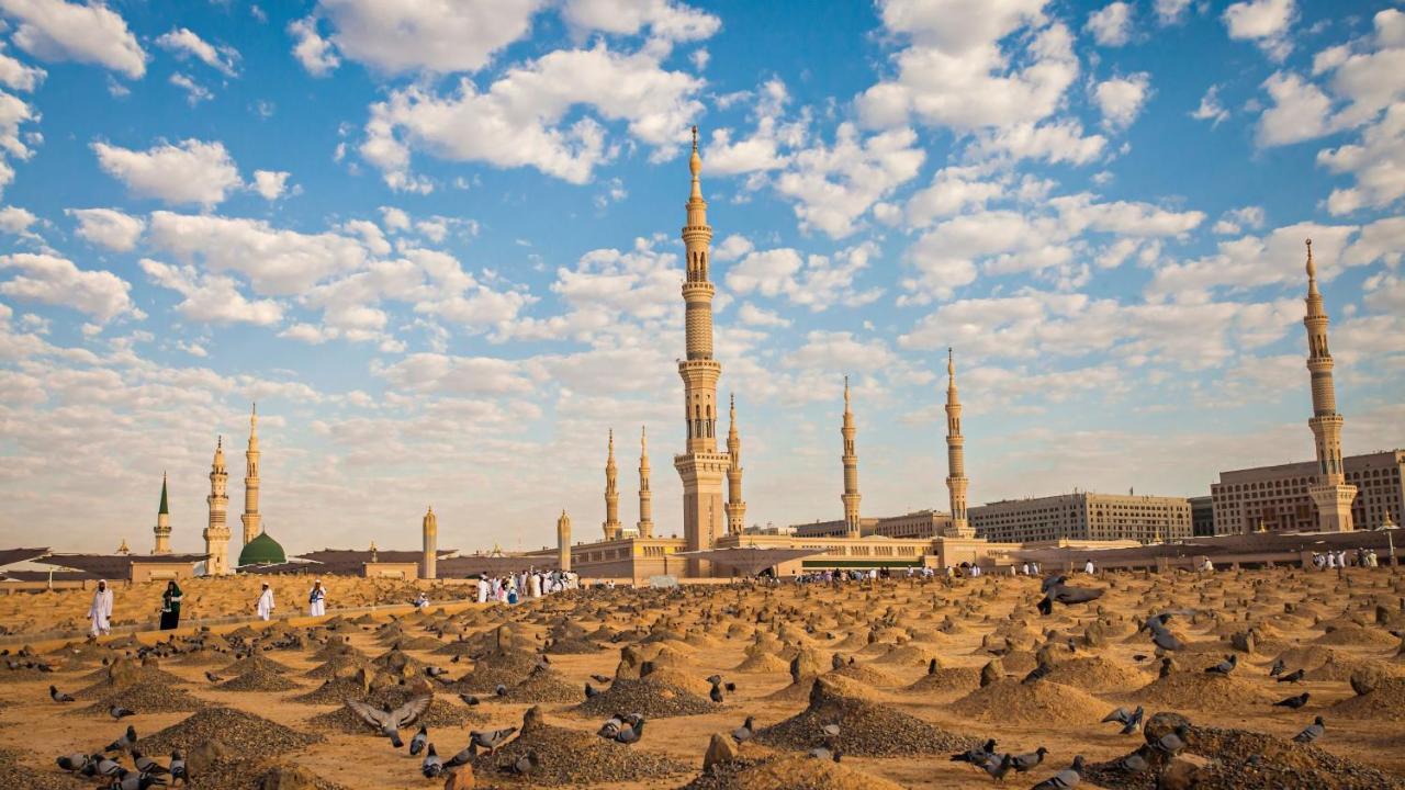 Intercontinental Dar Al Iman Madinah By Ihg Hotel Medina Exterior foto The photo shows a landscape that features a large mosque with a tall minaret surrounded by smaller minarets. In the foreground, there are numerous grave markers arranged in a desert-like setting. The sky is bright with scattered clouds, and there are