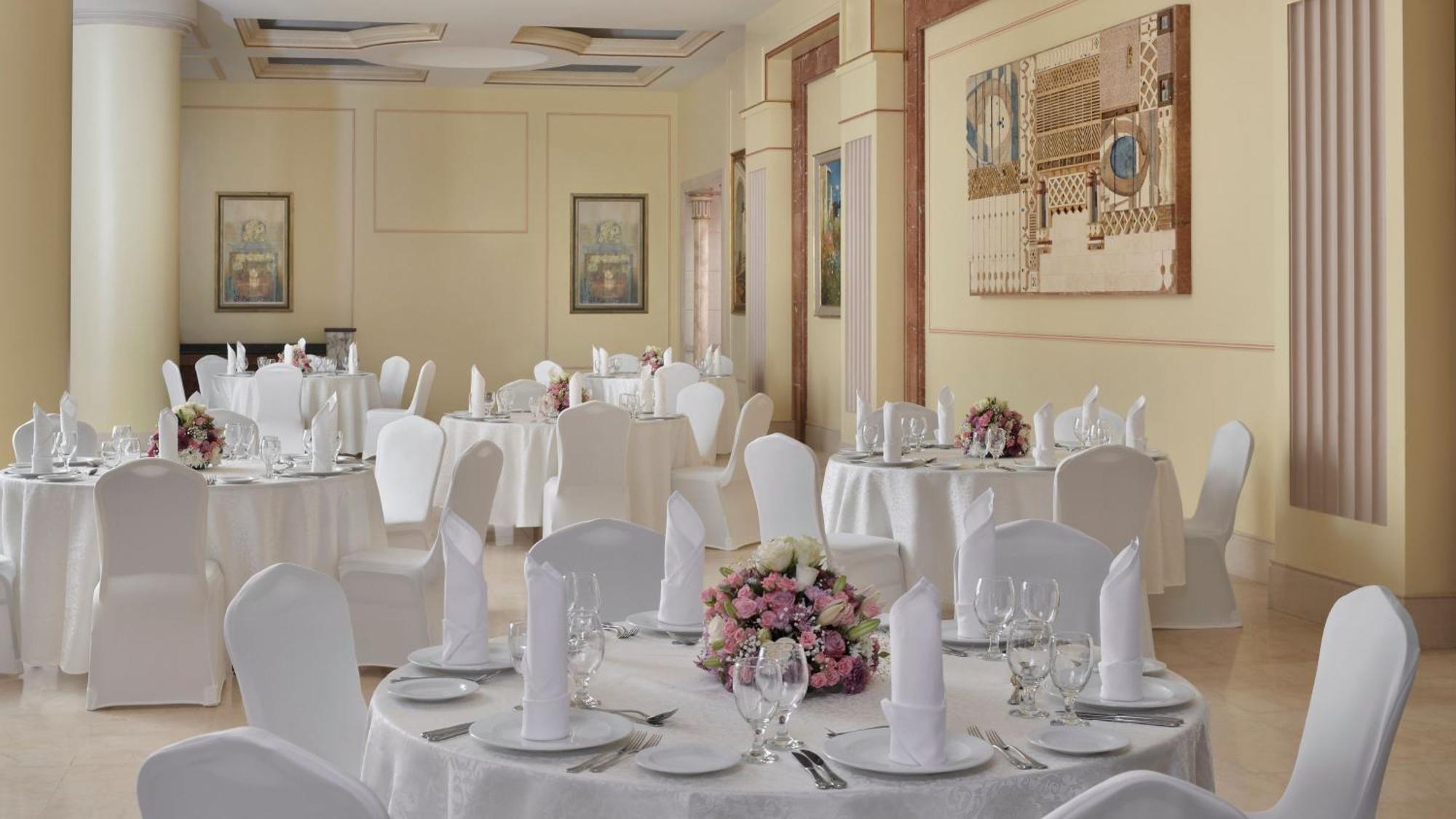 Intercontinental Dar Al Iman Madinah By Ihg Hotel Medina Exterior foto The photo shows an elegantly arranged dining area, likely set up for a formal event or reception. There are several round tables covered with white tablecloths, each adorned with floral centerpieces featuring pink flowers. The tables are set with pla