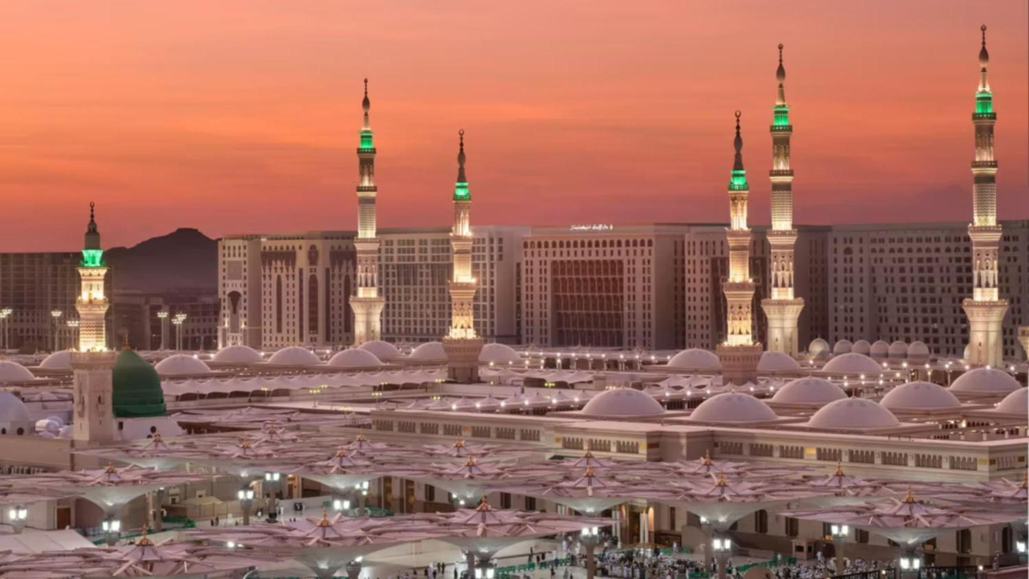 Intercontinental Dar Al Iman Madinah By Ihg Hotel Medina Exterior foto The photo depicts a scenic view of a mosque complex, likely during sunset, with several minarets illuminated and showcasing green lights. The architecture features domed roofs, and the setting includes a backdrop of buildings, all bathed in a warm, o