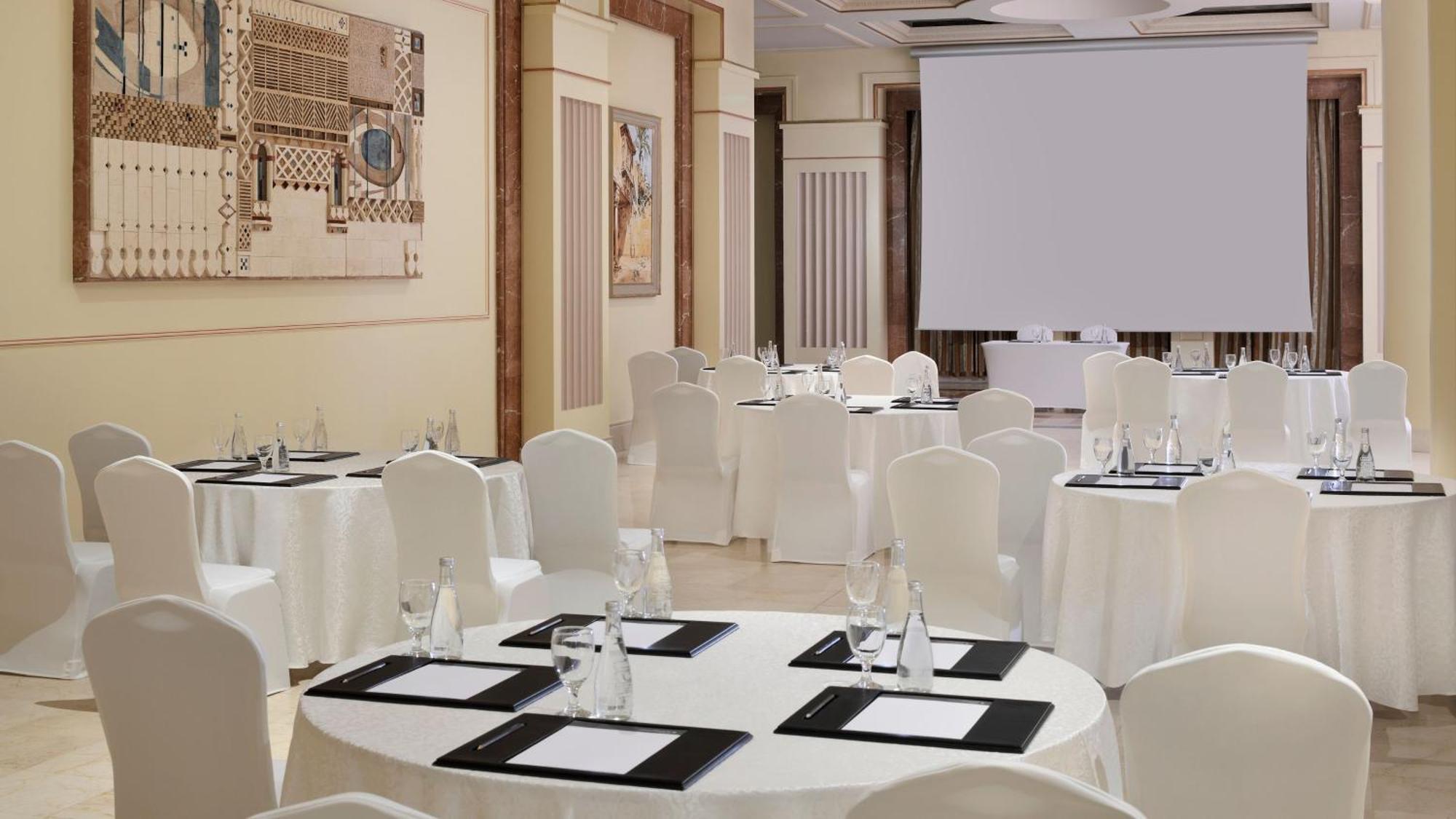 Intercontinental Dar Al Iman Madinah By Ihg Hotel Medina Exterior foto The photo depicts an elegantly arranged event space. There are several round tables covered with white tablecloths and surrounded by white chairs. Each table is set with a black folder or menu and a glass of water. The walls are adorned with artworks