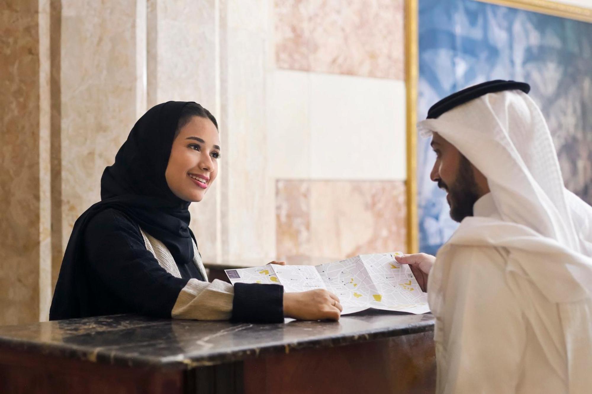 Intercontinental Dar Al Iman Madinah By Ihg Hotel Medina Exterior foto The photo depicts a woman in a black hijab and a light-colored outfit smiling as she interacts with a man wearing traditional Emirati attire, including a white kandura and ghutrah (headscarf). They are at a reception desk in what appears to be a hote