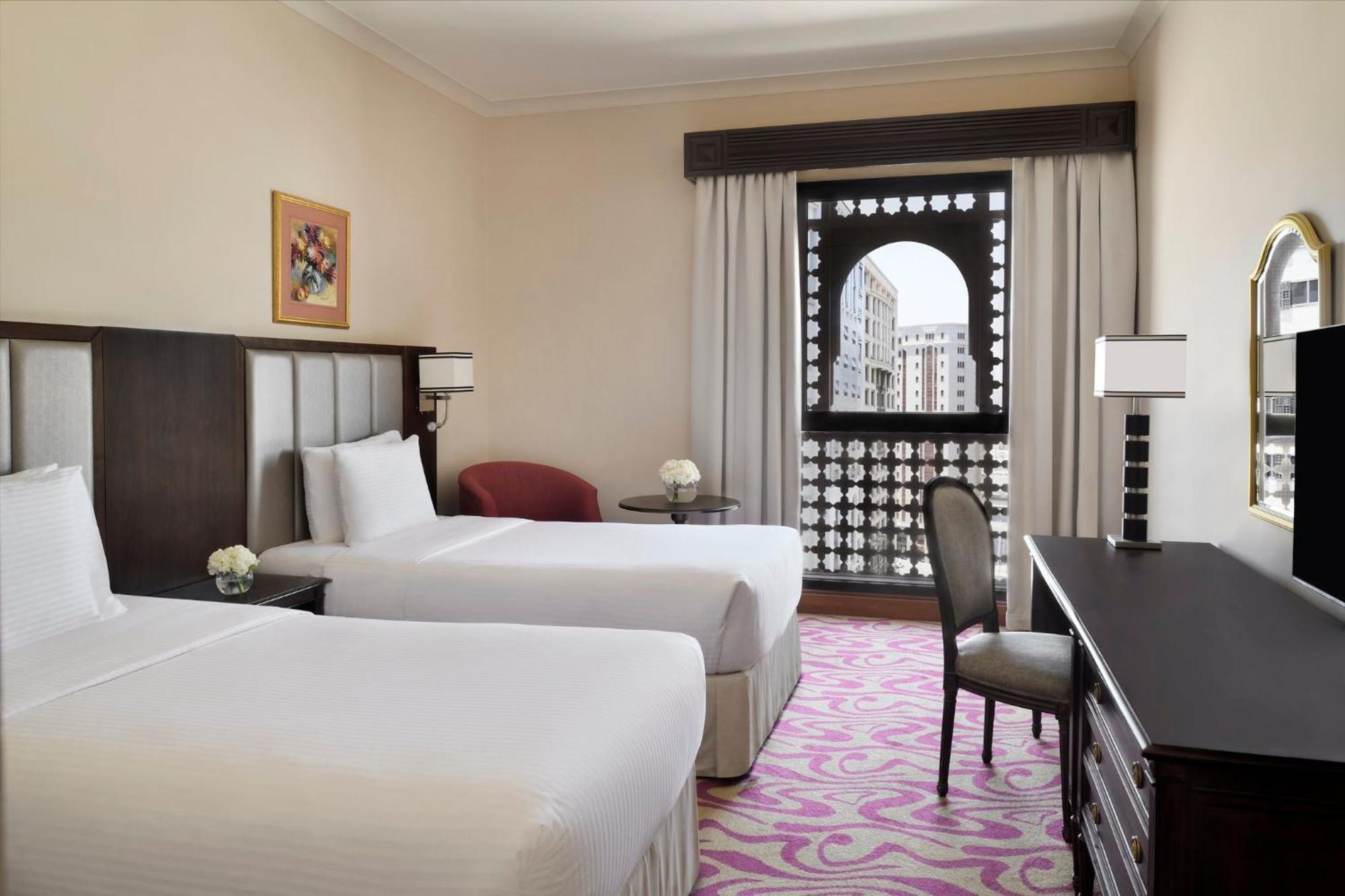 Intercontinental Dar Al Iman Madinah By Ihg Hotel Medina Exterior foto The photo shows a hotel room featuring two single beds with white bedding. The room has a light, neutral color scheme and is decorated with minimal furnishings. There's a small seating area with a red chair and a round table, as well as a desk with a
