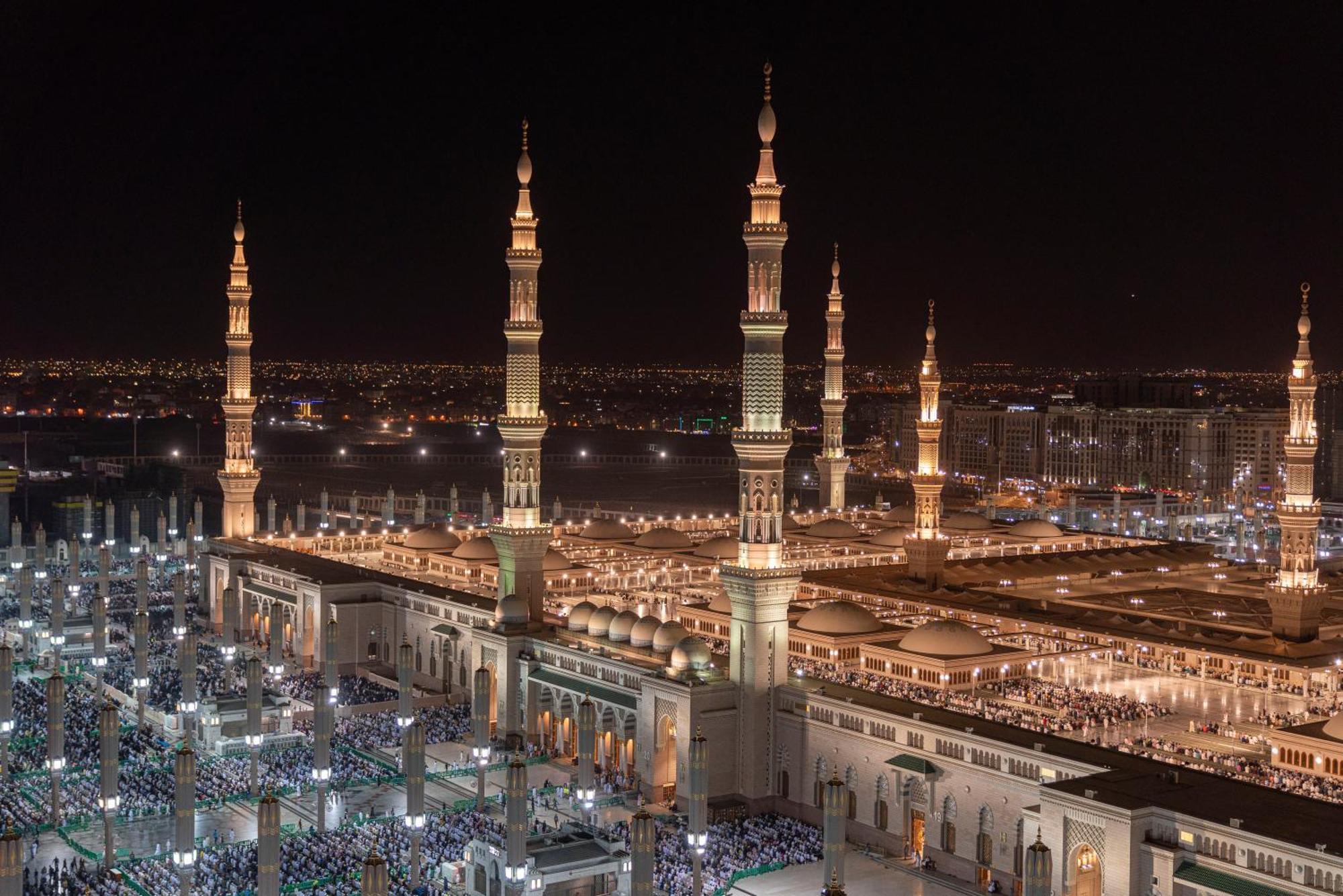 Intercontinental Dar Al Iman Madinah By Ihg Hotel Medina Exterior foto The photo depicts a stunning nighttime view of a large mosque complex, prominently featuring tall minarets that are illuminated against the dark sky. The surrounding area is bustling with people, likely worshippers, gathered in the courtyard of the m