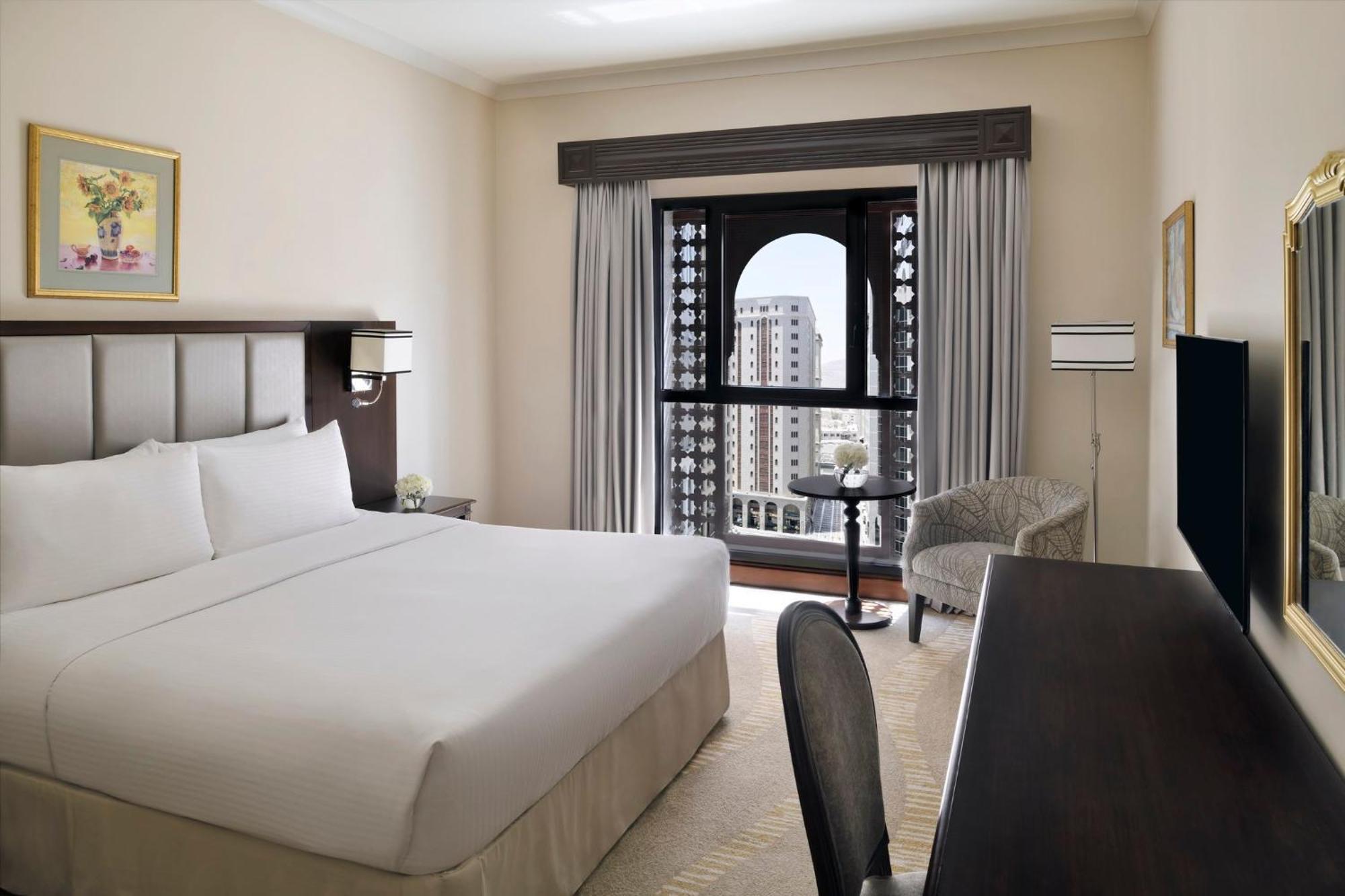 Intercontinental Dar Al Iman Madinah By Ihg Hotel Medina Exterior foto The photo shows a hotel room with a modern and minimalistic design. It features a large bed with white linens, a side table, and a comfortable chair near a window. The window has a view of a cityscape, and elegant curtains frame the window. There is 