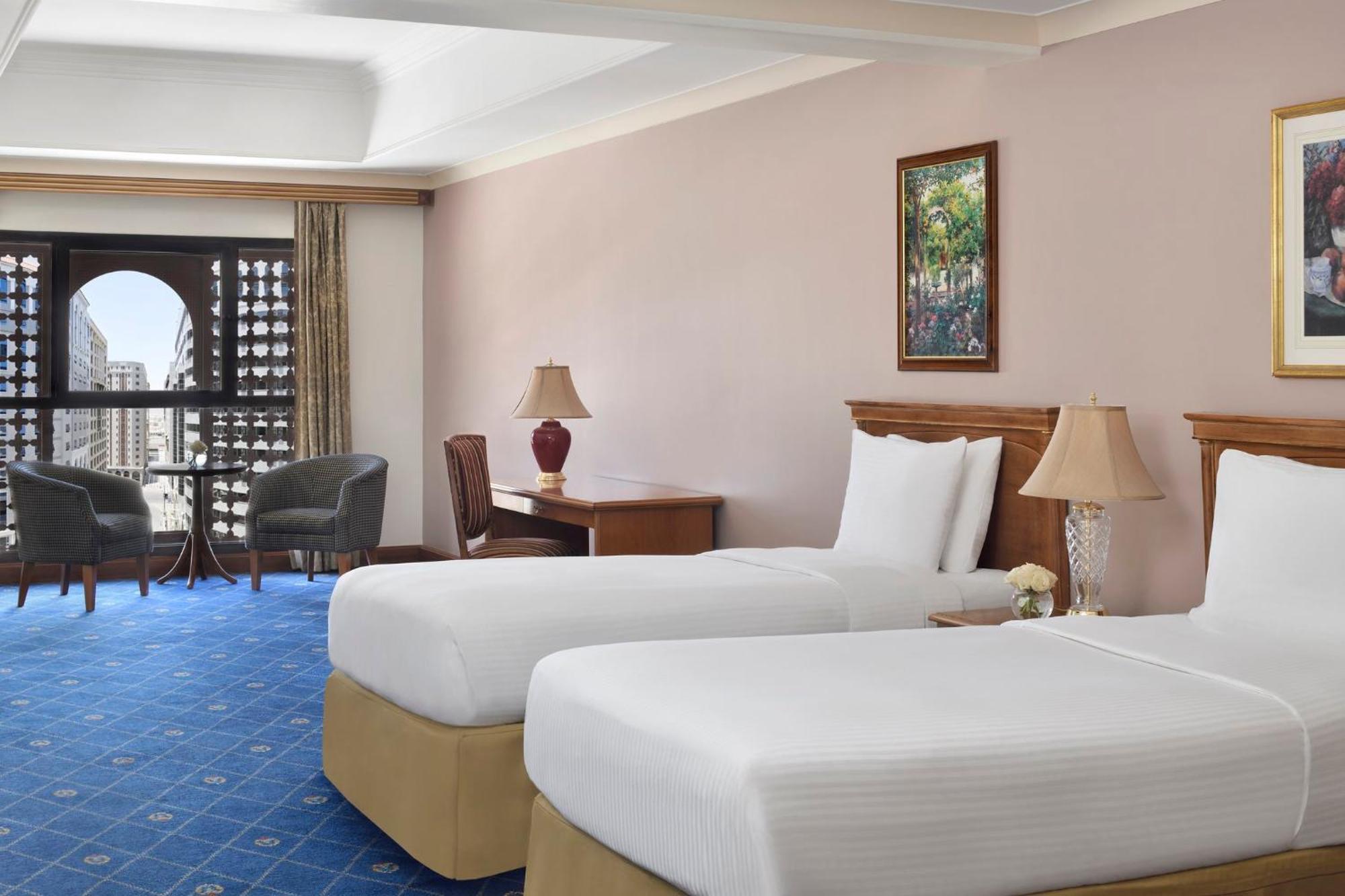 Intercontinental Dar Al Iman Madinah By Ihg Hotel Medina Exterior foto The photo shows a nicely designed hotel room featuring two twin beds with white linens and beige bases. In the background, there's a wooden desk and a lamp, along with a pair of comfortable armchairs positioned near a large window. The window showcas