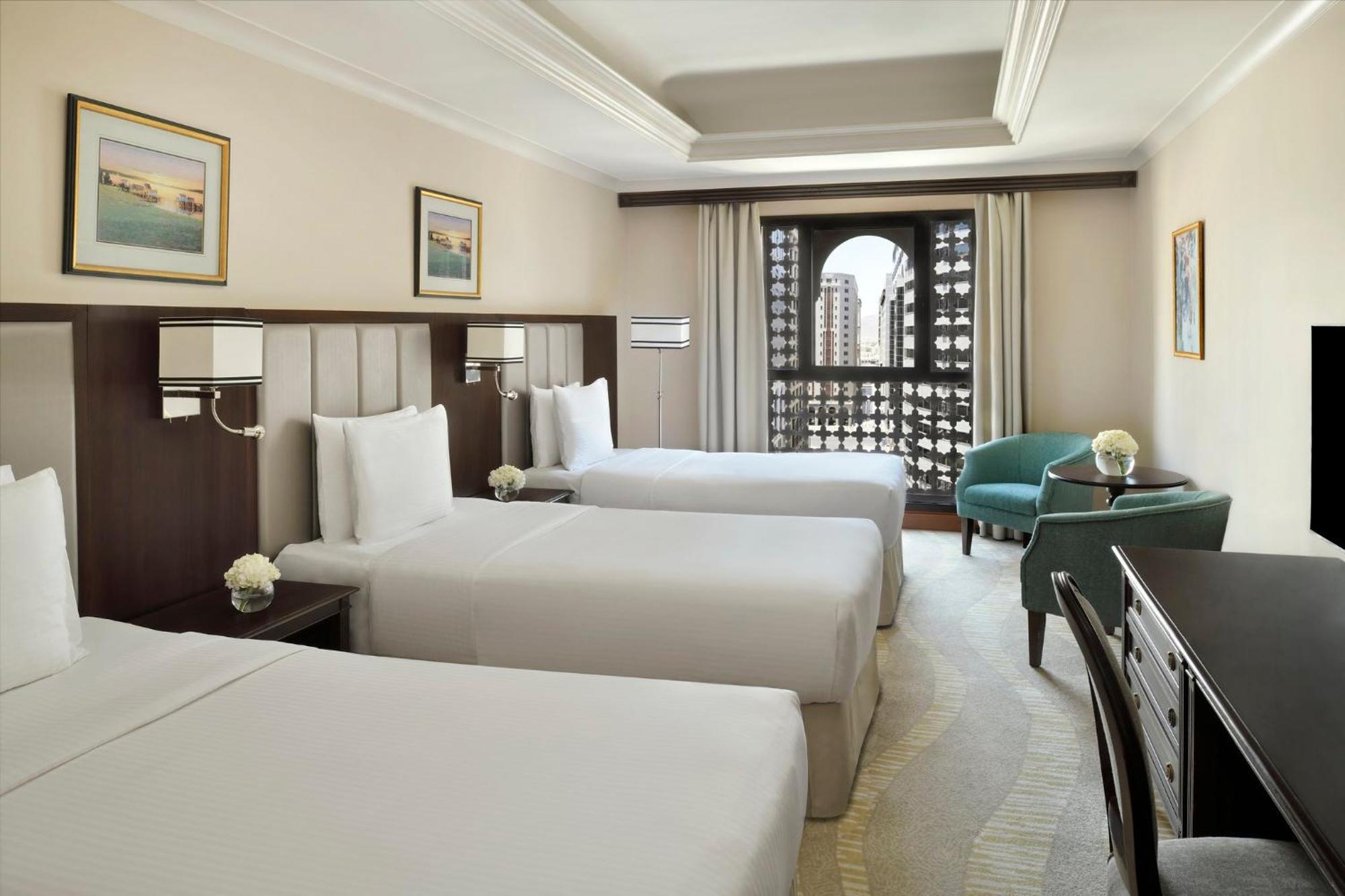 Intercontinental Dar Al Iman Madinah By Ihg Hotel Medina Exterior foto The photo shows a hotel room with three single beds, dressed in white linens. There are two paintings on the walls, depicting serene landscapes. A large window allows natural light to enter, and it is adorned with elegant curtains. A smaller seating 