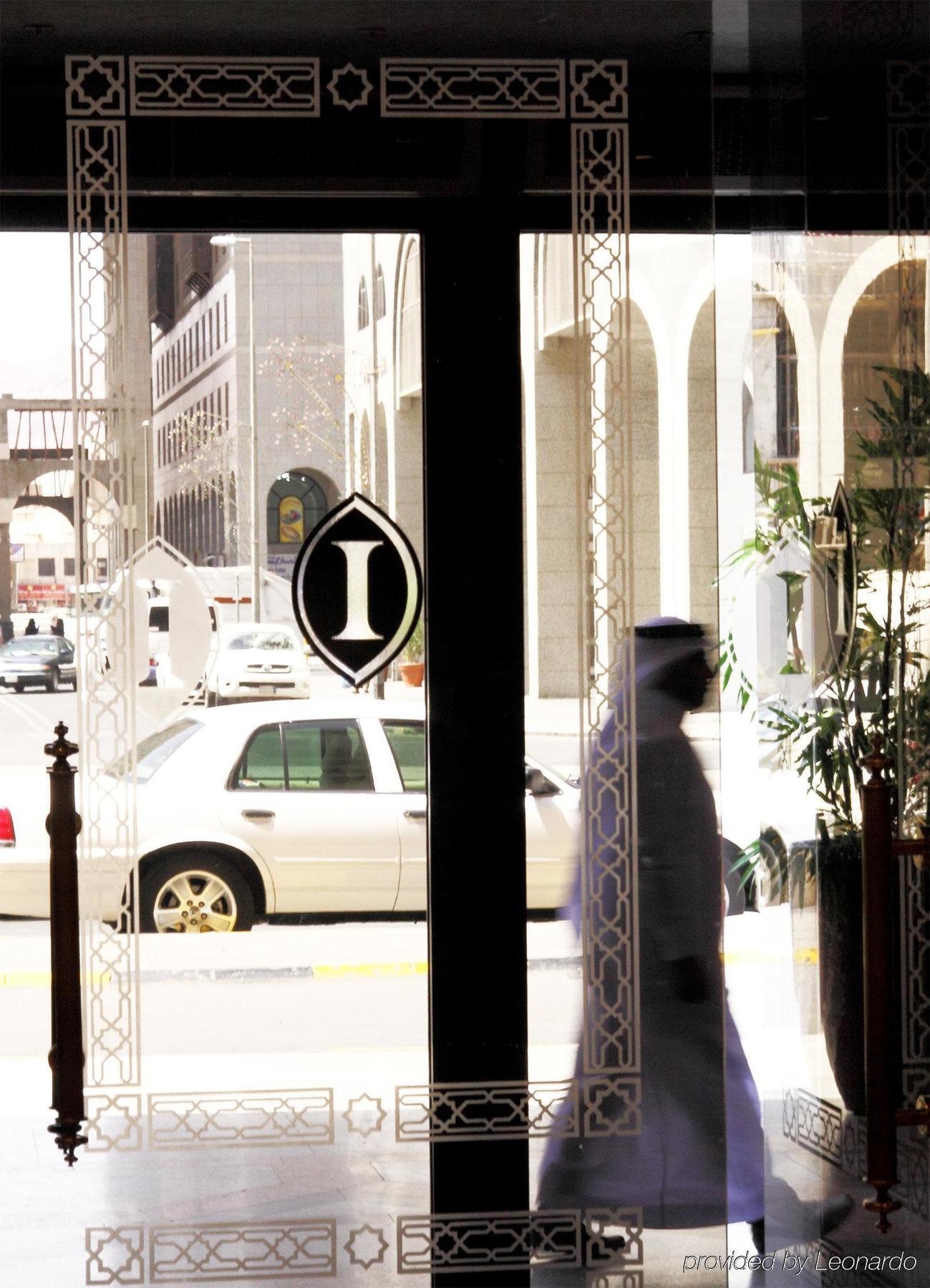 Intercontinental Dar Al Iman Madinah By Ihg Hotel Medina Interior foto The photo shows a view through a glass door or window, featuring an elegant design with a prominent "I" symbol displayed. Through the glass, there are some parked vehicles visible, including a white car. In the foreground, a shadowy figure of a perso
