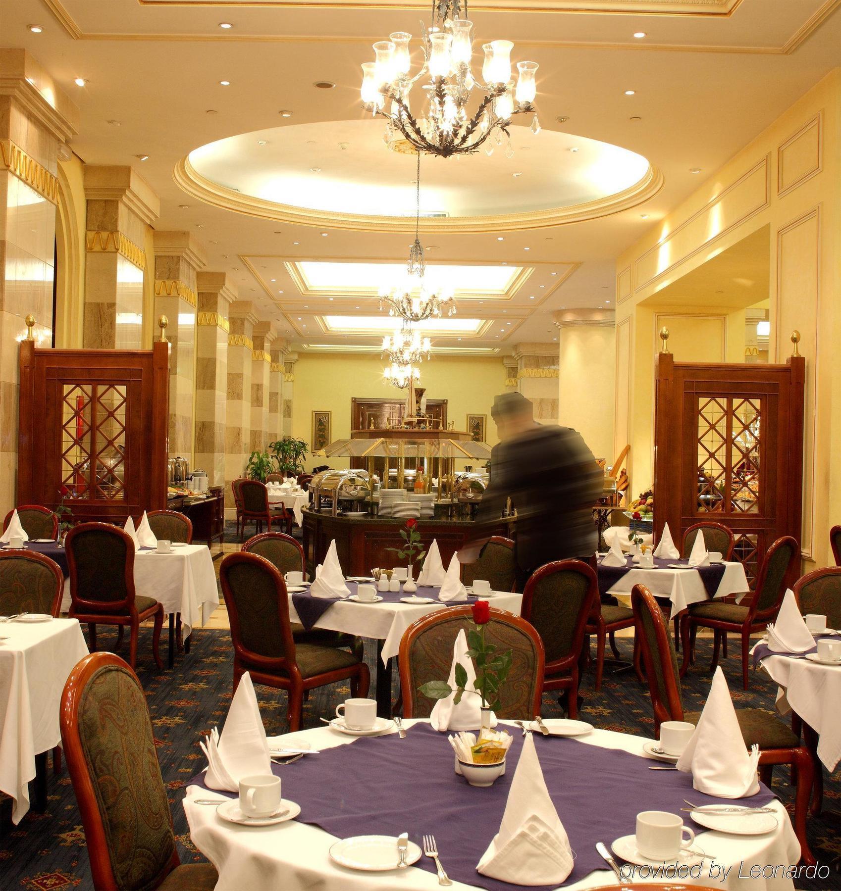 Intercontinental Dar Al Iman Madinah By Ihg Hotel Medina Restaurante foto The photo depicts an elegant restaurant interior. It features round tables covered with white tablecloths, each set with plates, cutlery, and folded napkins arranged into triangles. The space has a warm, inviting atmosphere with a mix of decorative e