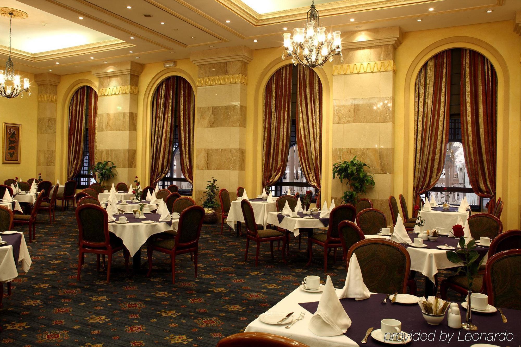 Intercontinental Dar Al Iman Madinah By Ihg Hotel Medina Restaurante foto The photo depicts an elegant restaurant interior. The space features ornate decor with tall windows draped in rich, patterned curtains. Tables are neatly set with white tablecloths and napkins, and there are plush chairs arranged around them. The flo